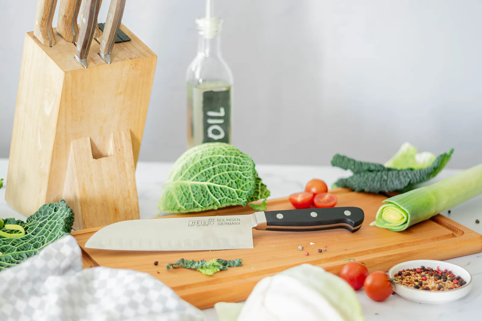 Stilvolle Food-Fotografie eines HEISO Solingen Santoku-Messers auf einem Schneidebrett, umgeben von frischem Gemüse wie Wirsing, Lauch und Tomaten, inszeniert von Socialfood.