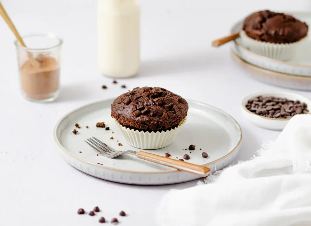 Food-Fotografie von SOCIALFOOD: Schokoladen-Muffin mit Schokostückchen, stilvoll inszeniert auf einem weißen Teller mit Besteck und dezenten Accessoires im Hintergrund.