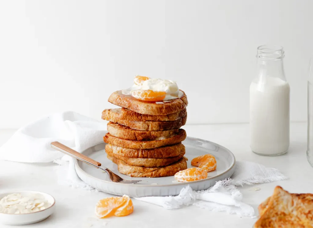Food-Styling von SOCIALFOOD: Kunstvoll gestapeltes French Toast mit Mandarinen und Sahne auf einem weißen Teller, inszeniert mit Milchflaschen und feinen Details im Hintergrund.