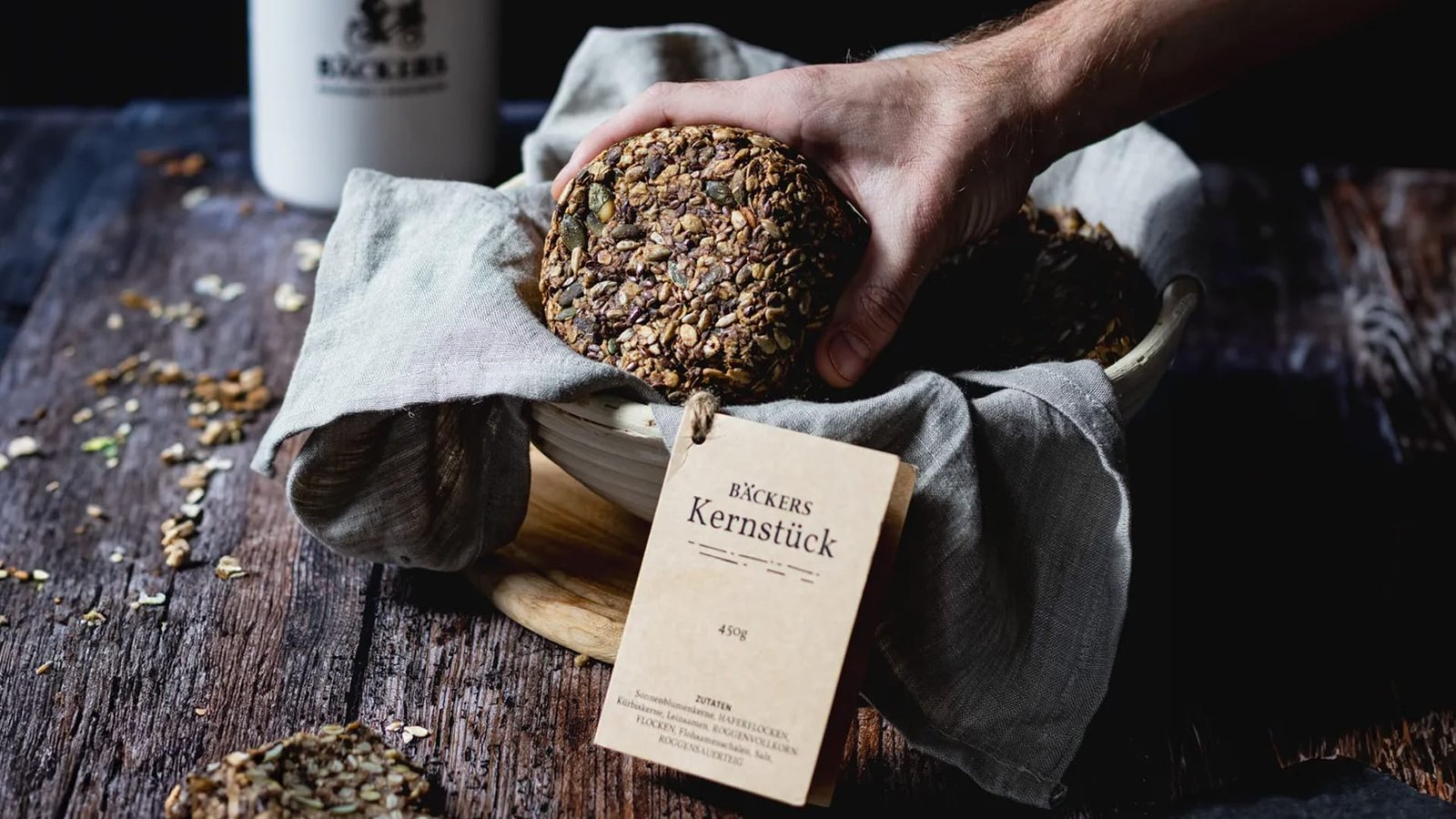 Food-Fotografie in Wien – Hand legt frisches Bäckers Kernstück mit Körnerkruste in eine rustikale Schale, stilvoll inszeniert auf Holzhintergrund.
